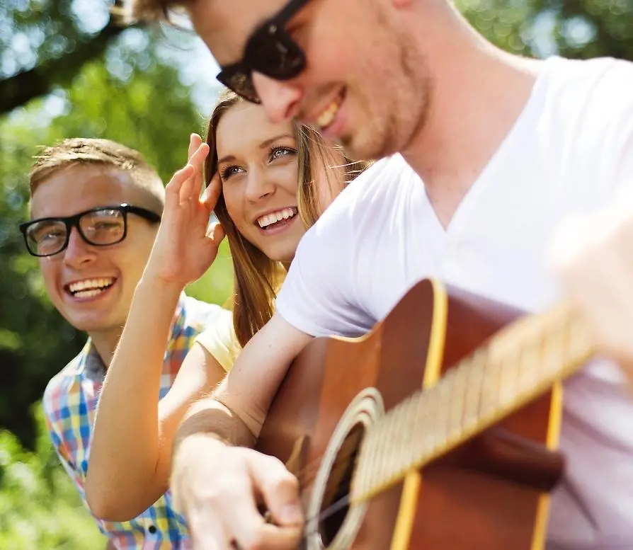 Tre ungdomar som ser glada ut tillsammans. De är utomhus i sommartid och en av dem spelar gitarr.