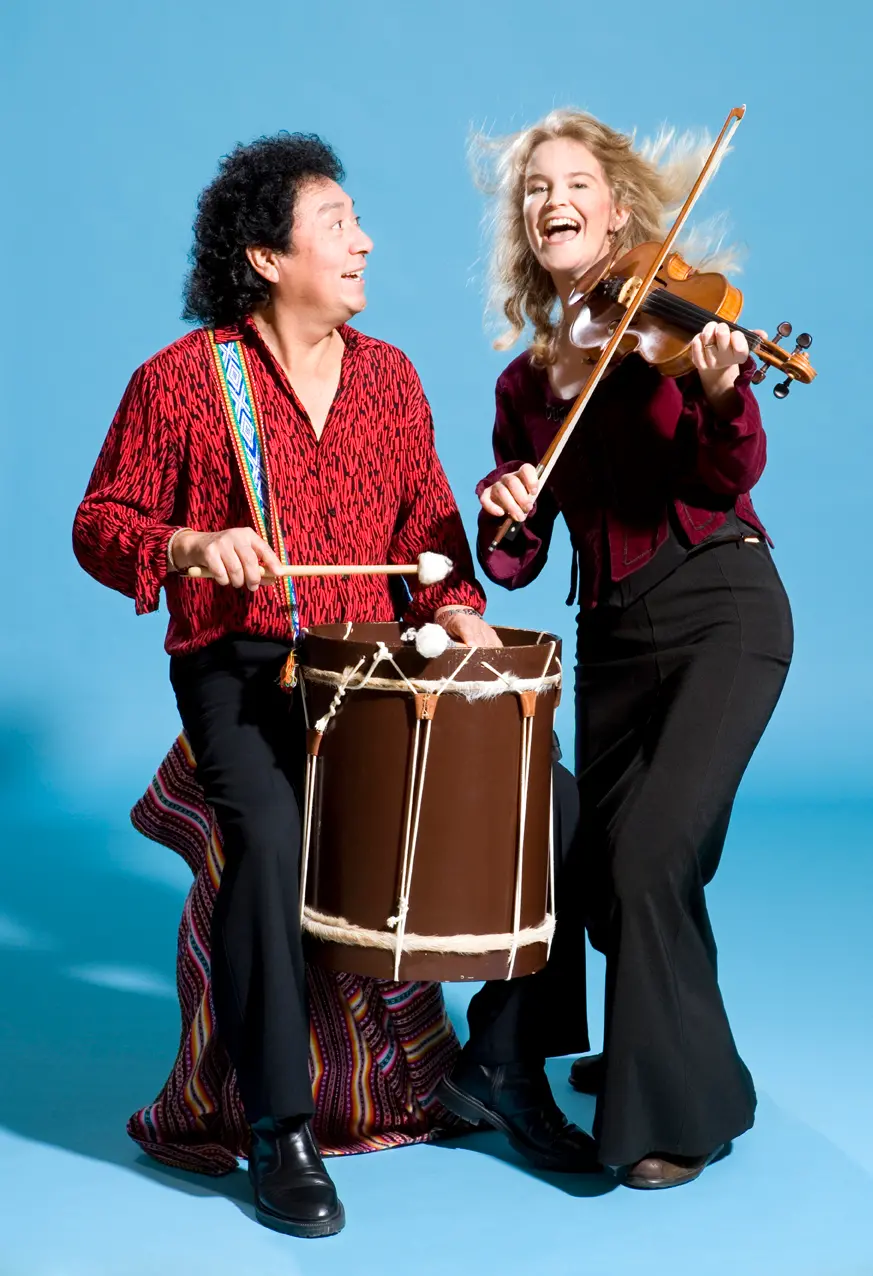 Två glada personer mot en klarblå bakgrund. Till vänster en med röd skjorta, svarta byxor, och en puka framför sig, som hänger över högra axeln. Till höger en glad fiolspelare, också med röd skjorta och svarta byxor.