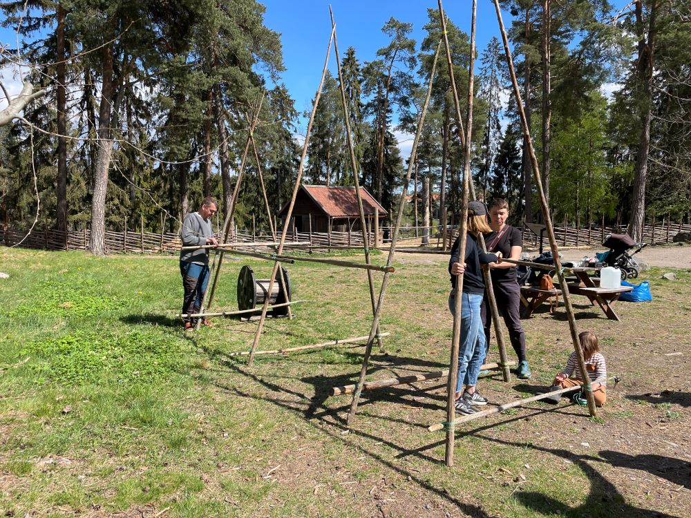 Tre personer bygger koja med långa pinnar på Vallby friluftsmuseum.