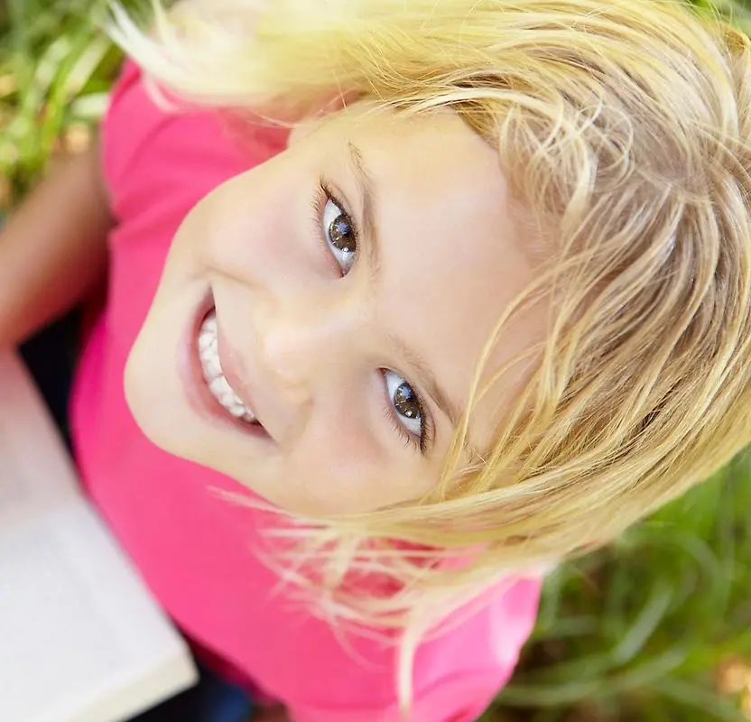 En flicka med blont hår och rosa tröja sitter i gräset och läser en bok. Bilden är tagen ovanifrån och flickan tittar rakt upp mot kameran med ett leende.
