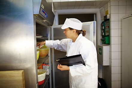 På bilden sker en livsmedelskontroll på en restaurang av en kvinna i vit rock och skyddsmössa.