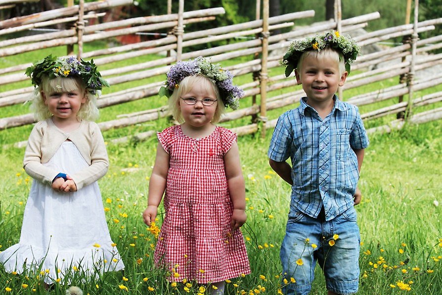 Tre barn står framför en gärdesgård och är klädda i sommarkläder och har en blomsterkrans på huvudet.  
